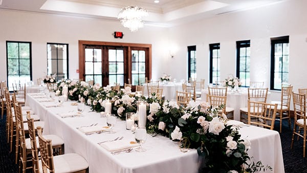 Elegant indoor wedding reception setup at Gervasi Vineyard with white linens, lush floral arrangements, and gold chairs.