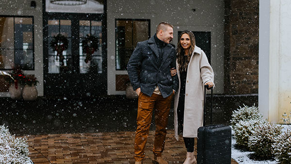 Smiling couple arriving at Gervasi Vineyard during light snowfall, dressed in winter coats and ready for a cozy seasonal getaway