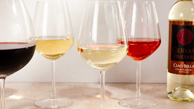 Close-up of five wine glasses filled with Gervasi Vineyard’s red, white, and rosé wines, including a visible bottle of Ciao Bella Chardonnay, showcasing varietal colors and textures for warm-weather enjoyment.