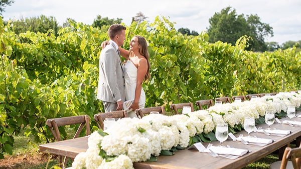 Newlyweds embrace in front of a vineyard at Gervasi Vineyard, beside a long rustic table adorned with white hydrangeas and elegant place settings.