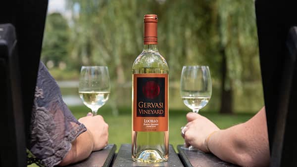 Two guests relaxing by the lake with glasses of Lucello white wine at Gervasi Vineyard’s scenic outdoor tasting area.