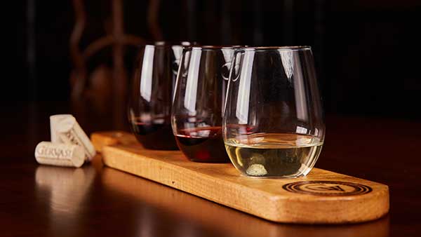 A wooden flight board holding three glasses of red and white wine beside several Gervasi-branded corks, displayed on a dark wood table.